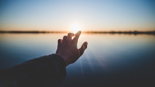 man's outreached finger touching sunsetting sun over water